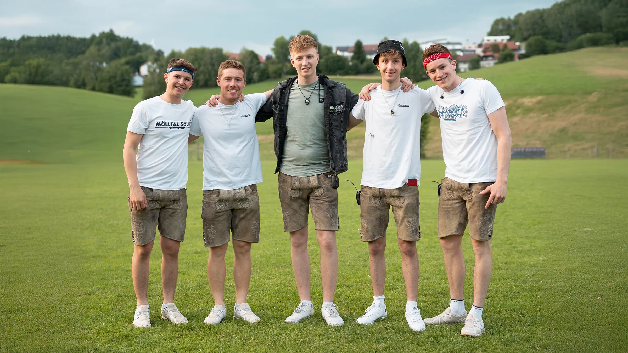 Die fünf Mitglieder von MölltalSound stehen nebeneinander auf einer grünen Wiese und lächeln in die Kamera. Von links nach rechts: Florian Kranabether, Fabian Angerer, René Zlöbl, Marco Zlöbl und Elias Wallner. Die Bandmitglieder tragen weiße T-Shirts mit MölltalSound-Aufdruck sowie kurze Lederhosen und weiße Sneakers.